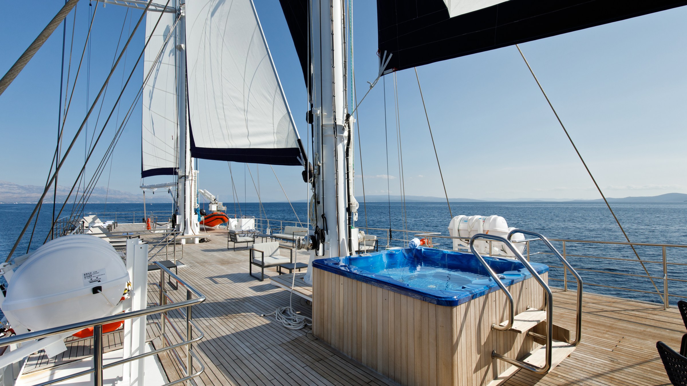 Sailboat deck with sails, hot tub, and ocean view under clear sky.