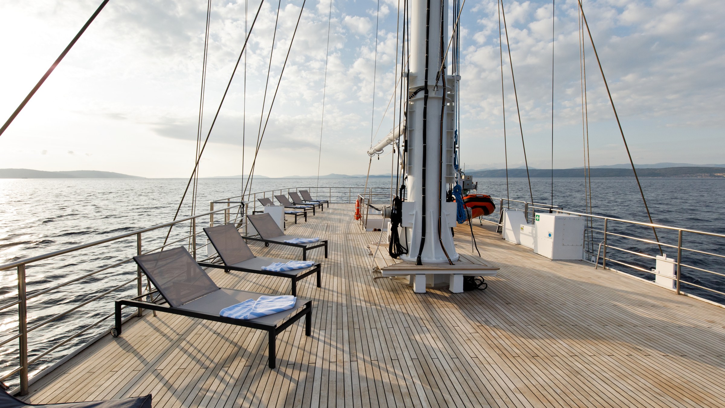 Deck of a yacht with empty loungers and ocean view under a partly cloudy sky.