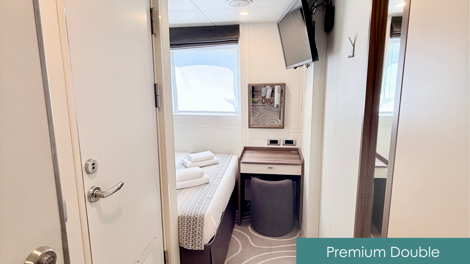 Double cabin room with a bed, TV, and desk. Towels on bed. Mirror and window behind.