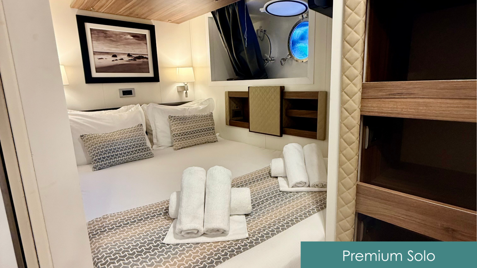 Compact bedroom with a porthole, neatly arranged bed, and towels, labeled 'Premium Solo'.