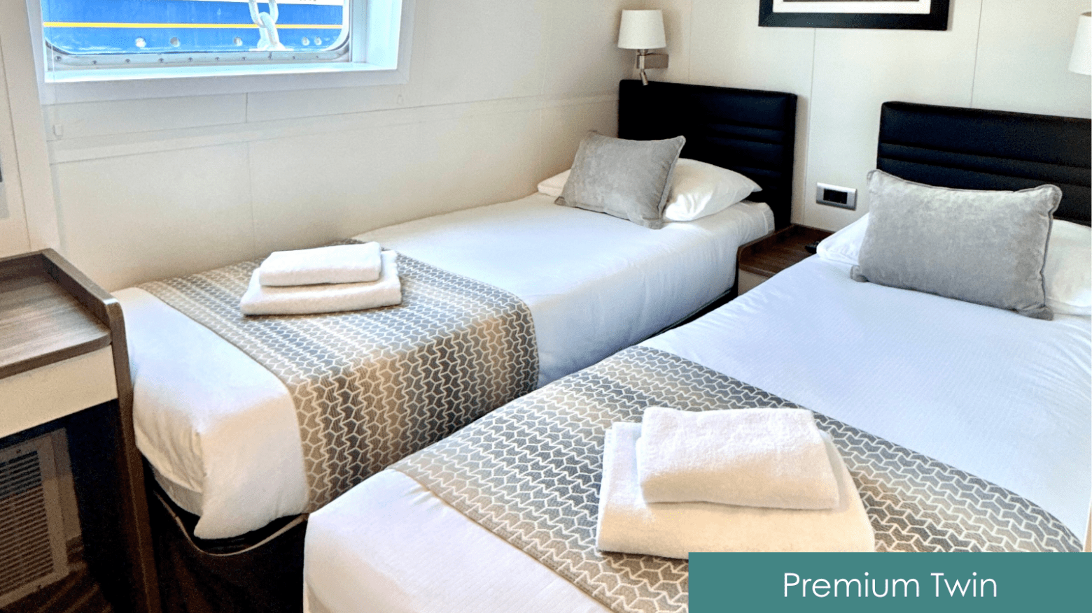 Two twin beds with gray pillows and patterned runners, neatly made with towels, under a window in a bright room.