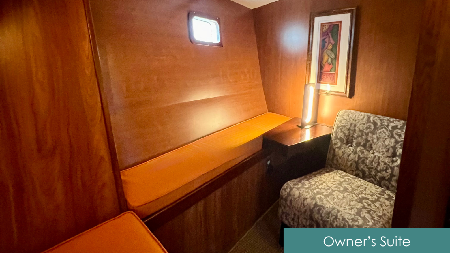 Wood-paneled room with an orange cushioned bench, patterned chair, and artwork on the wall.
