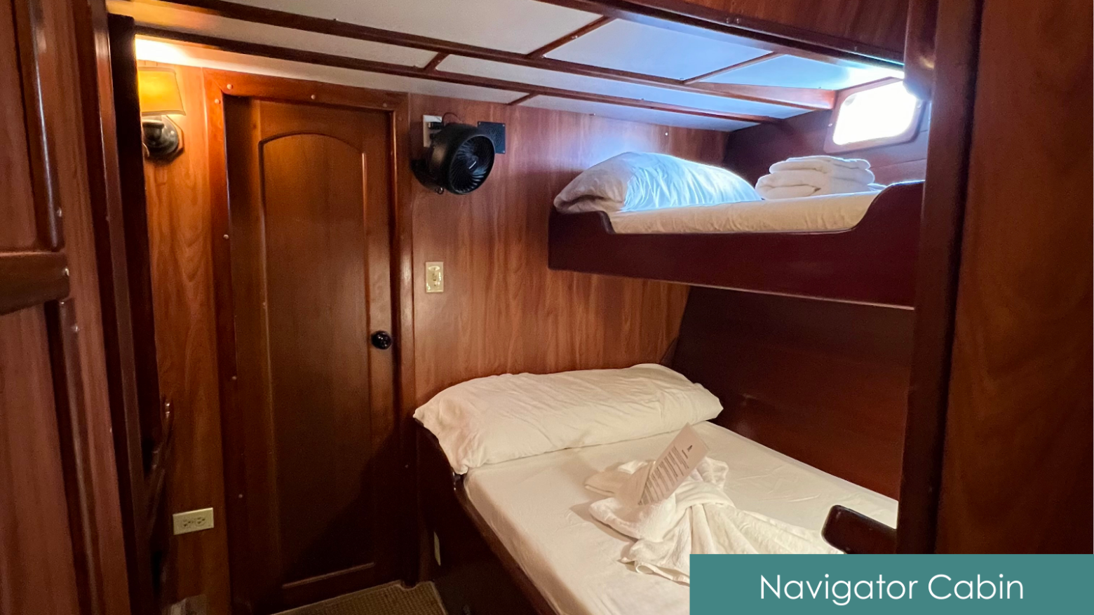 Cozy wooden cabin with bunk beds, fan, and small window, labeled 'Navigator Cabin'.