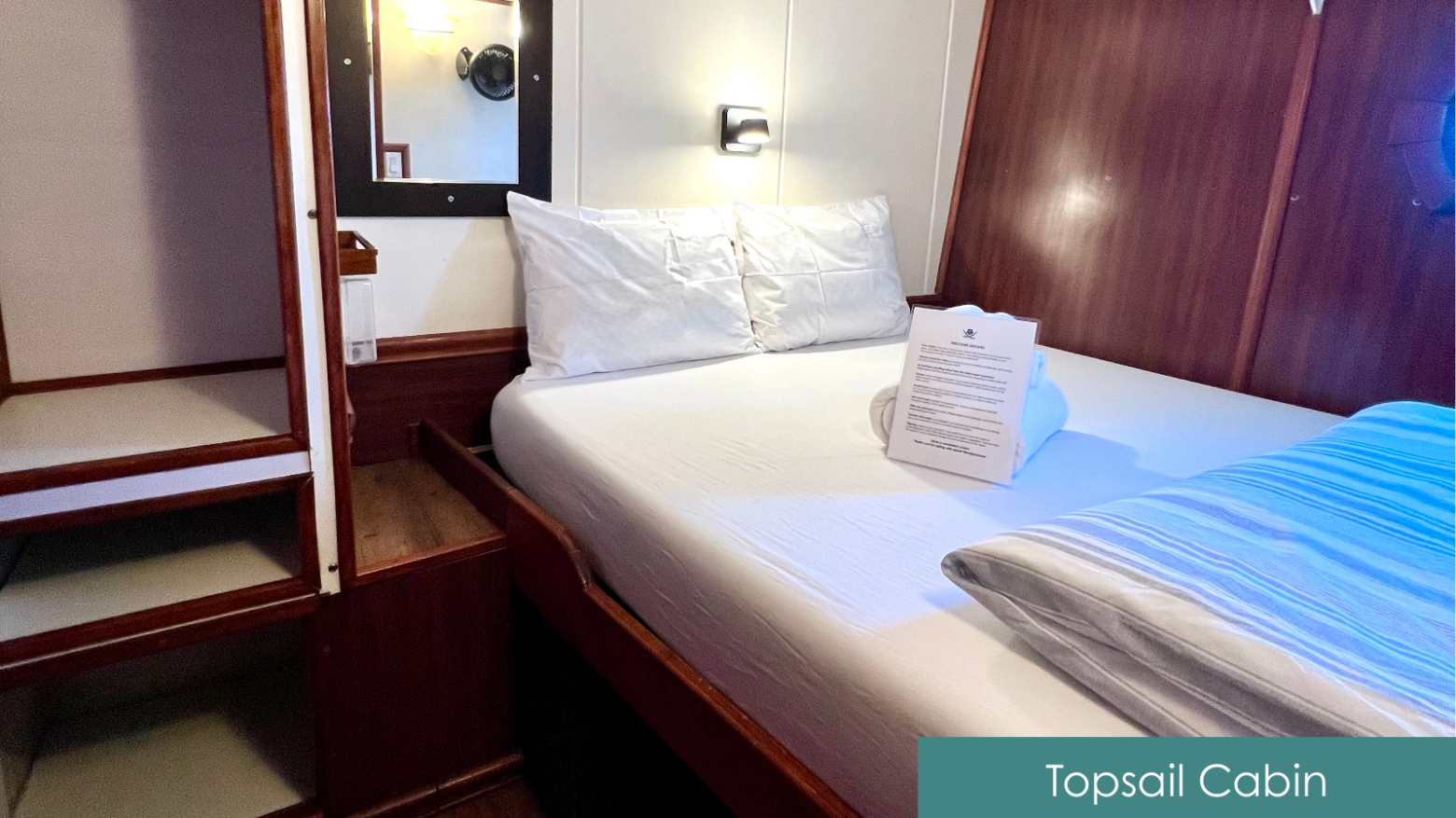 Cozy boat cabin with a double bed, white linens, and a porthole window. Sign reads 'Topsail Cabin'.