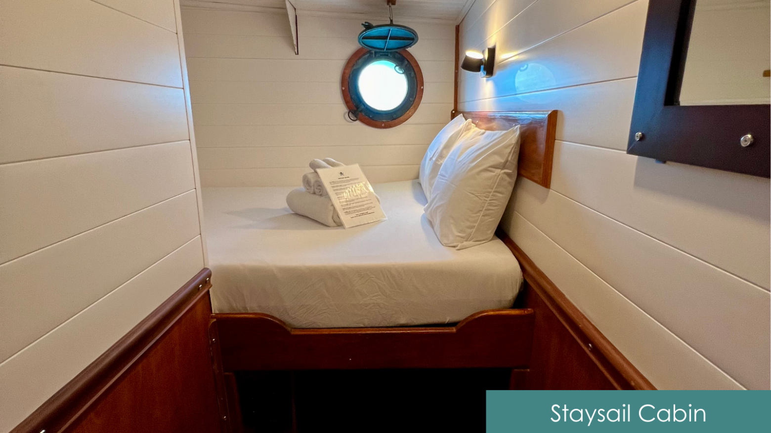 Small ship cabin with round porthole, a bed, and folded towels.