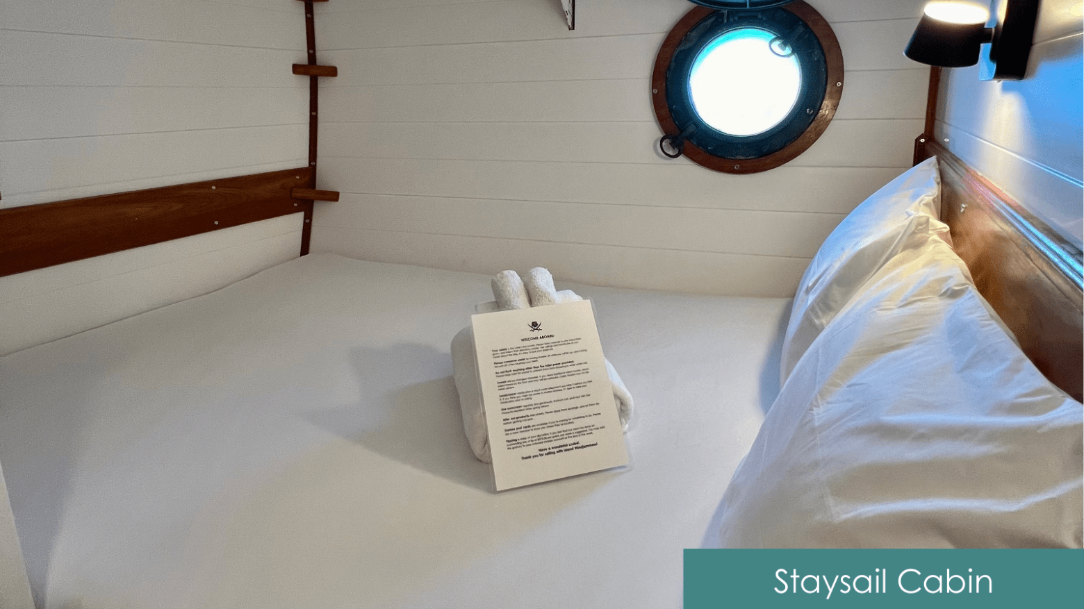 Cabin bed with porthole, folded towels, and welcome note on bed.