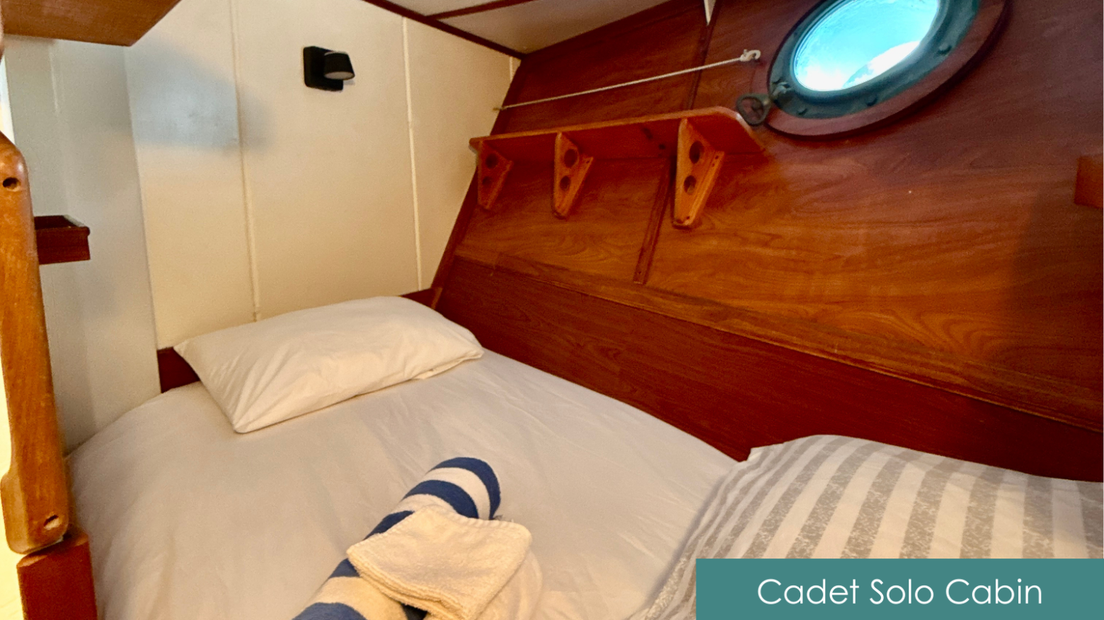 Wooden cabin with a single bed, porthole, and striped towel on the bed.