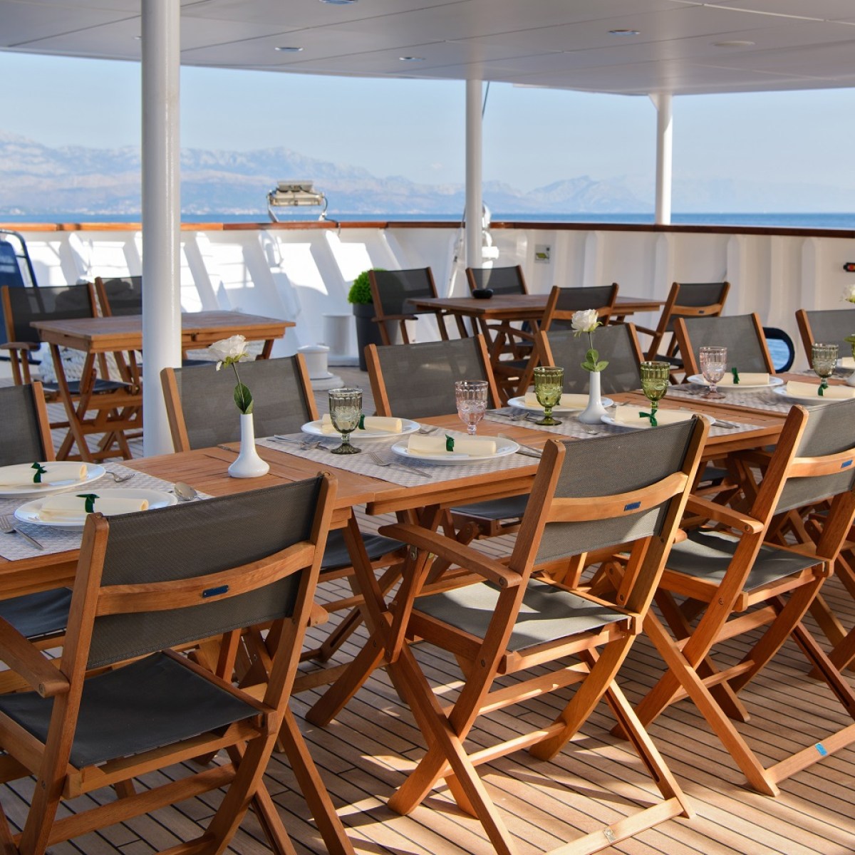 Outdoor dining area on a ship with wooden tables and chairs, set for a meal.
