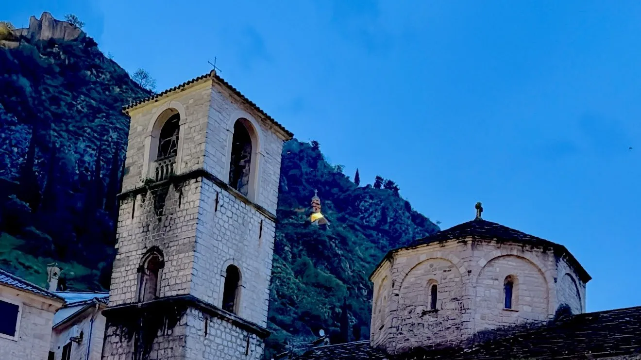 Stone fountain in courtyard with old church and mountain backdrop at dusk.