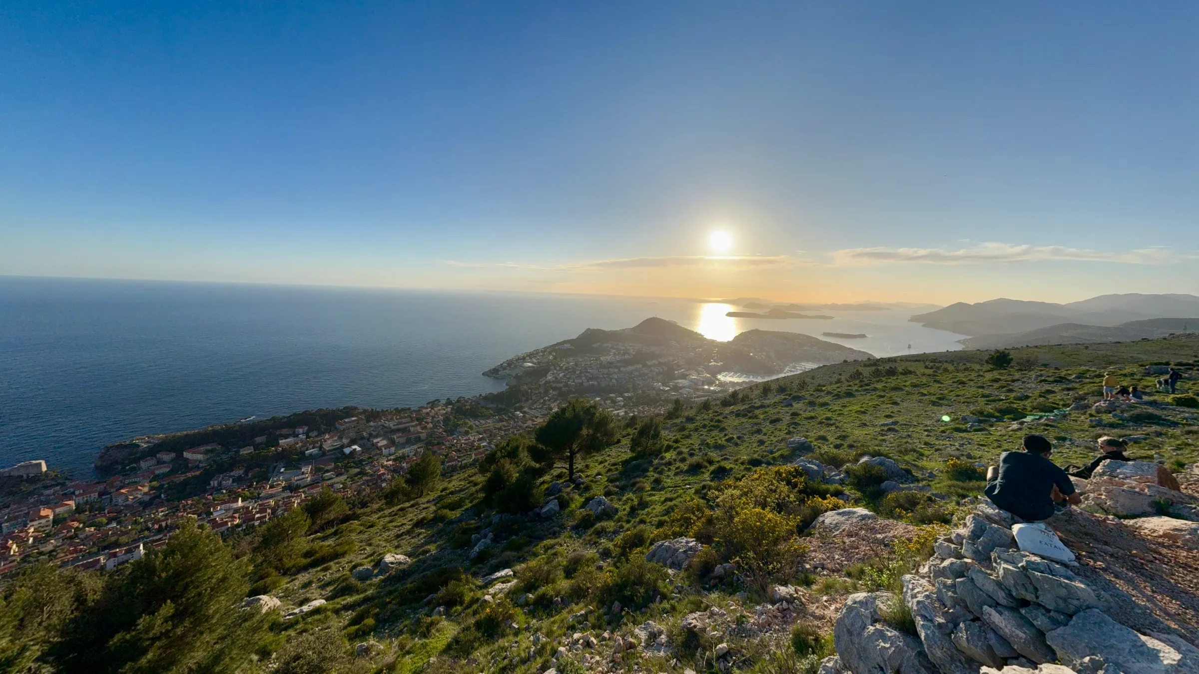Scenic coastal sunset view from a rocky hill with a person sitting and distant islands.