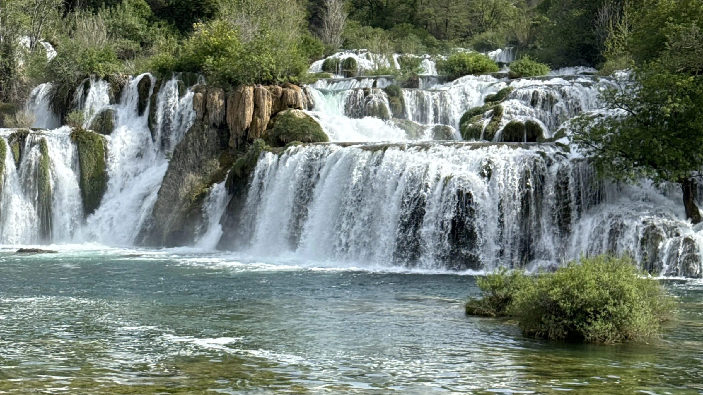 Waterfall cascading over rocks with greenery and clear water.