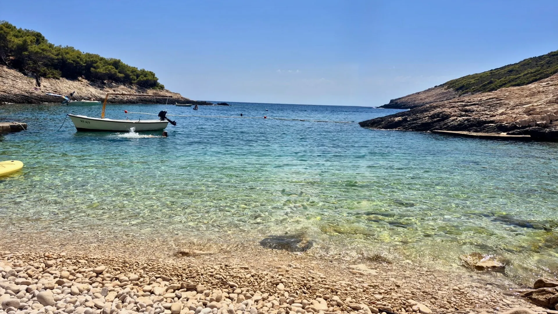 Pebble beach with a small boat on clear water between two rocky hills under a blue sky.